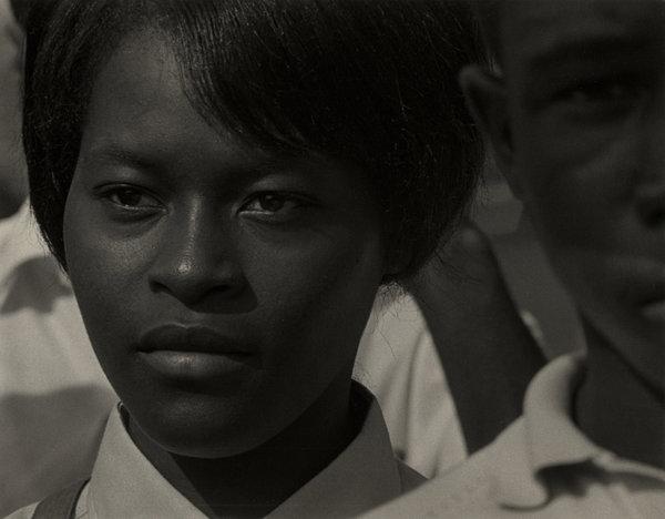 Fotografía en blanco y negro de una joven negra vestida de uniforme, de pie entre otras personas negras.
